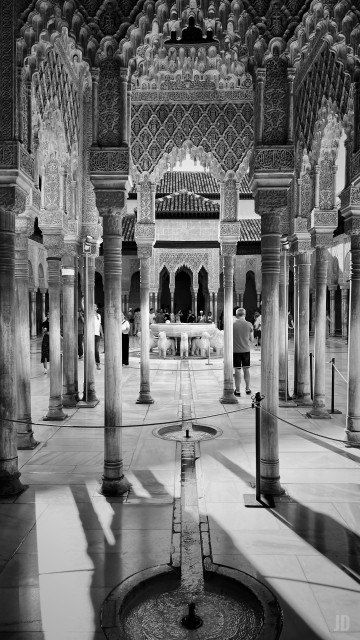 La imagen muestra el Patio de Los Leones, en La Alhambra de Granada, de arquitectura islámica con columnas esbeltas y arcos decorados con intrincados relieves geométricos y caligráficos. En el centro se aprecia una fuente rodeada por el conjunto de leones esculpidos, mientras unos visitantes caminan y observan el lugar. El suelo está pavimentado con piedra y un canal de agua conecta varias pequeñas fuentes circulares, reflejando la luz y las sombras que atraviesan el espacio.