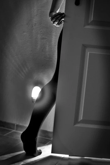 Eine Frau geht durch eine Tür. Nur die rechte Hand ist zu sehen, die die Tür festhält, und das rechte Bein, nur in einer Strumpfhose gekleidet. Durch die leicht geöffnete Tür fällt Licht in den Raum. An der Wand trägt ein kleines Nachtlicht zur spärlichen Beleuchtung bei. 