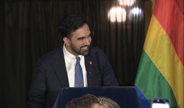 A still image from a video of New York Mayor Zohran Mamdani speaking, smiling at a podium next to a rainbow pride flag. 