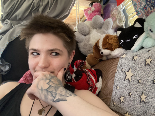 A person with short hair is resting their chin on their hand, looking thoughtfully. Behind them, various plush toys are arranged, including a large gray bunny on its back, a King Charles spaniel dog, and a few other colorful characters. The setting appears cozy and bright with blankets