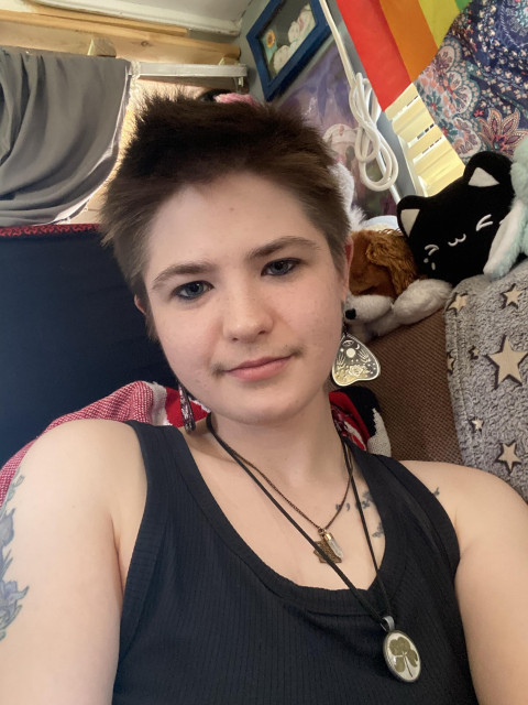 A person with short hair is sitting in a cozy space surrounded by colorful decor and plush toys. They are wearing a black tank top and multiple necklaces, featuring earrings and visible tattoos on their arm. The background includes various items like a rainbow flag and patterned
