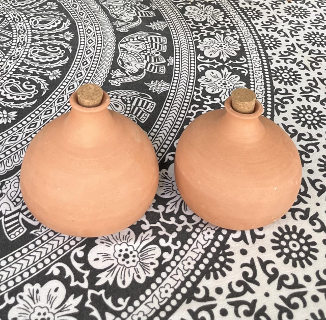 Two fat-bellied Terra Cotta pots with short narrow neck with cork stopper plugging the hole. They are sitting on a black and white Indian print tablecloth.