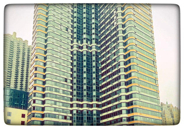 A color photo of a huge tower of condos, probably built in the 1960s or 70s. It consists of a tower on each side that is larger and a thin tower in the middle. Each level of windows is separated by a large strip of color. In this case I have brought the natural oranges and greens up a bit and the windows look black in several places. There are also three smaller buildings on the left and one at the corner on the right. The photo has been made to look very futuristic. And there is a frame on it that looks like a TV screen.