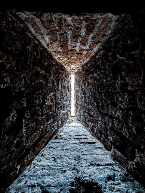 A high-contrast perspective looking through a narrow, stone-walled loophole toward a bright opening.

The stone floor, walls, and ceiling converge sharply toward the center of the frame, creating a powerful sense of depth and a tunnel vision effect.

The walls and floor are constructed from heavily textured, uneven blocks of stone or brick, characteristic of ancient fortifications.