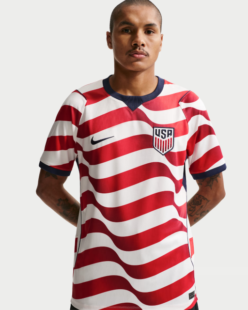A man poses facing the camera with his hands behind his back, wearing a red-and-white horizontally striped USA soccer jersey with a navy collar and sleeve trim. The jersey features a Nike logo on one side and a USA crest on the other. He has a shaved head, a mustache, and visible arm tattoos.
