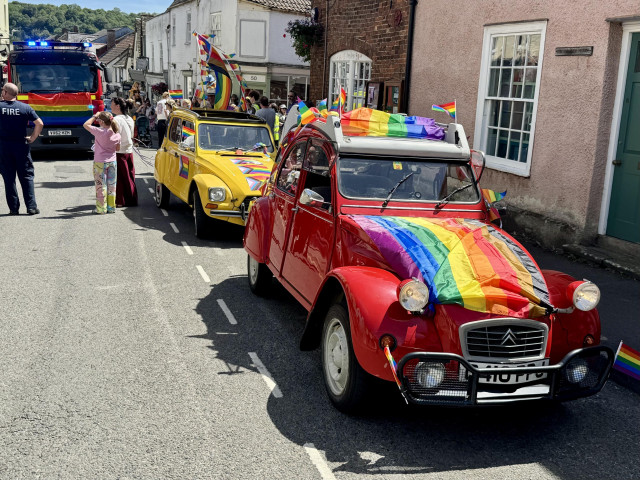 Two old Citroen 2CVs, red and yellow, and a fire engine, all with various Pride flags.