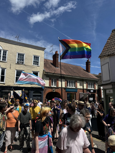 Photo or the parade passing down a Cotswold high street against a blue sky with Pride progress and Trans Pride flags held high.