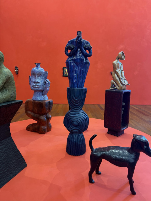 An art installation featuring several sculptures with a vibrant red wall in the background. The sculptures vary in shape and style, including human-like forms and an abstract dog figure, arranged on a circular platform. 