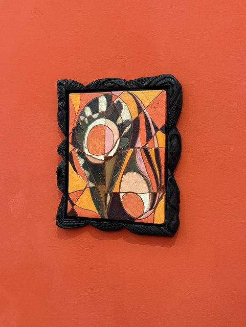 An abstract ceramic mosaic artwork framed in ornate black wood, featuring geometric shapes and vibrant colors like orange, green, and brown. The background is a solid red-orange wall.