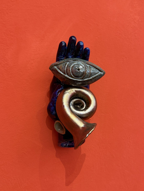 A sculpture featuring a hand in a dark blue color, an eye, and a curled horn shape. The background is a vibrant red-orange wall.