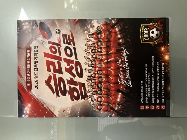 A promotional poster is taped to a glass wall. The design is dominated by red, white, and black colours and depicts a large soccer stadium filled with lights and cheering fans. A South Korean flag and a soccer ball appear near the top right, reinforcing the World Cup theme.

Large Korean text in the centre reads “Victory Anthem” (Song of Victory), with smaller text above announcing the 19th regular concert of the Vancouver Choir and describing it as a special 2026 World Cup performance.

At the bottom, approximately forty choir members pose for a group photograph. Most of the women wear matching red dresses, while several men wear white shirts with red bow ties. Many are raising clenched fists in a celebratory gesture.

An English slogan below the group reads: “Together as One! One Voice, One Victory!”

Event details near the bottom list:

* June 27, 2026, at 7:00 PM
* Chandos Pattison Auditorium, Surrey, British Columbia

Additional Korean text identifies sponsors and partners, including the Korean Consulate in Vancouver, a Korean veterans association, Hanin Credit Union, and GreenTee Country Club. The poster also advertises a prize draw for a Korean Air round-trip ticket to Korea.

A shield-shaped logo in the lower-right corner reads “Vancouver Korean Choir” and incorporates a soccer ball motif. Reflections of people and nearby lights are faintly visible in the glass behind the poster.