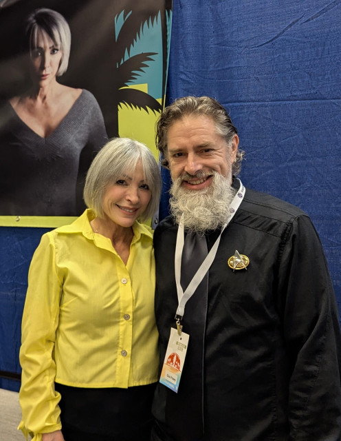 Nana Visitor from film and television, especially Deep Space Nine, standing next to me, a tallish white guy with curly hair and a thick gray beard.  In this example I'm wearing a black shirt and a dark gray tie, as well as my convention badge and a TNG-era communicator.