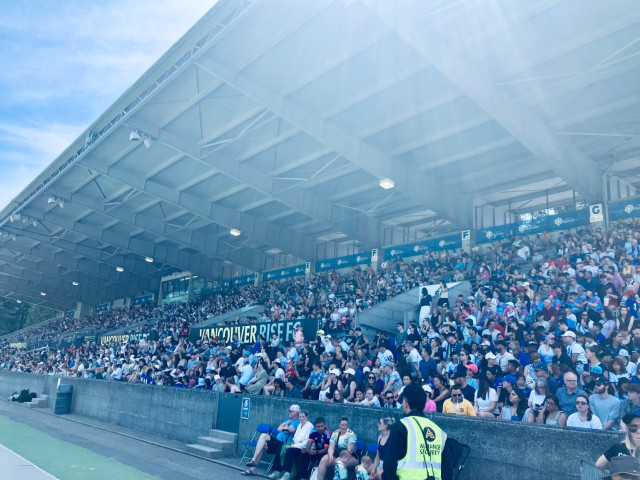 A large, roofed stadium grandstand packed with spectators on a bright sunny day. Hundreds of people fill the concrete bleachers, many wearing hats and sunglasses to shield themselves from the sun. A large sign reading “VANCOUVER RISE FC” stretches across the middle of the stands, indicating a soccer match or related sporting event. Sun glare and lens flare wash across the upper-right portion of the image, creating a hazy effect beneath the steel roof structure. In the foreground, a security staff member wearing a high-visibility yellow vest stands near the front row, while fans sit and chat along the lower seating area. The atmosphere appears crowded, warm, and energetic.