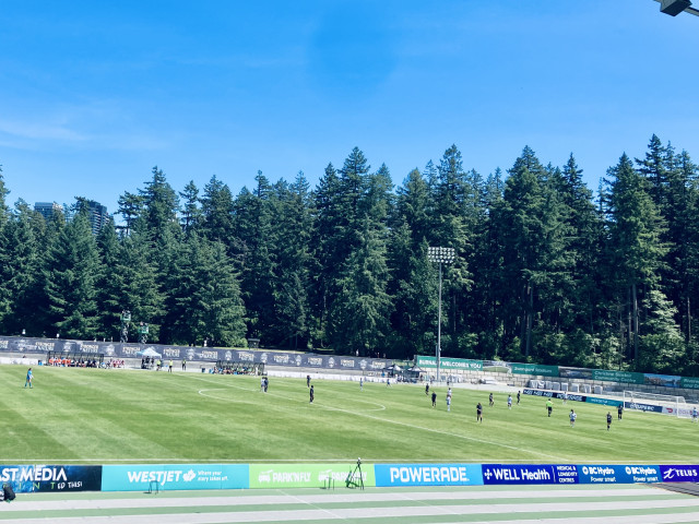 A wide view of an outdoor soccer match in progress at a stadium on a bright, clear day. Players in dark and light uniforms are spread across a well-maintained green pitch. Beyond the field stands a dense wall of tall evergreen trees, creating a forested backdrop that towers over the stadium. Advertising boards line the track surrounding the field, featuring brands such as WestJet, Powerade, TELUS, BC Hydro, and WELL Health. Stadium signage includes “Burnaby Welcomes You,” “Swangard Stadium,” and references to the Christine Sinclair Community Centre. A few high-rise buildings are visible peeking above the trees on the left side of the image. The sky is vivid blue with only faint wisps of cloud.