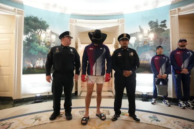 Foto von Al Drago. Eine Situation in einem ausgeleuchteten ovalen Raum. Eine Person in Sportkleidung, Cowboyhut und Badelatschen blickt auf den Boden. Die Person steht zwischen zwei Sicherheitsleuten in Uniform. Im Hintergrund stehen zwei weitere Personen in Trainingsanzügen, die sich gegen die Wand lehnen. Einer der beiden hält einen Eimer in seinen Händen.