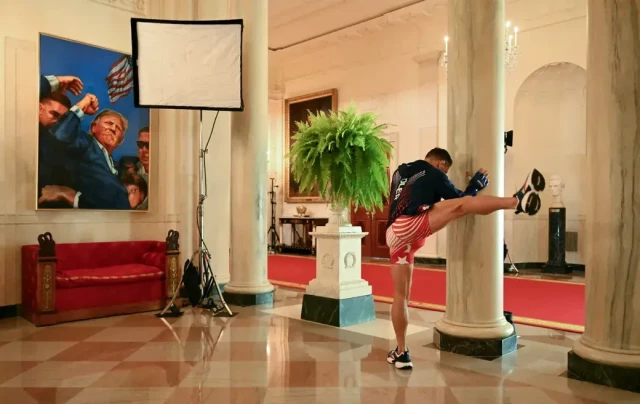 oto von Saul Loeb. Ein Sportler lehnt an einer Säule. Er steht mit dem Rücken zu uns und wärmt sich gerade auf, wobei er sein rechts bein hebt. das Ganze spielt sich in einem Bereich des Weißen Hauses ab. Man sieht im Hintergrund einen mit rotem Teppich ausgelegten Flur, Bilder an der Wand, eine Büste und einen Farn auf einer Stele. Oberhalb eies roten Sofas hängt ein Bild Donald Trumps, wie er die Faust reckt.