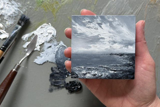 Photo of a hand holding an original seascape oil painting by Tisha Mark, "Tiny Respite" oil on cradled panel, 3"x3" (2026). This seascape painting is monochromatic (black & white). It has a sea on the left meeting a shore on the right. There is much texture throughout the painting, and there are moody cloud formations in the sky. A paint palette with puddles of oil paint, paintbrushes, and a palette knife are visible in the background.