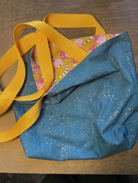 Photo of tote bag. The outer fabric is greenish blue with splatters of gold, black and purple. The lining fabric is a deep orange with pink flowers. The straps are a very bright gold webbing. 