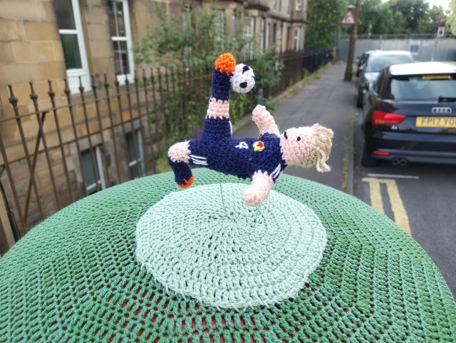 Knitted Scottish footballer McTominay, in mid air (on thin green wires), performing overheard kick of ball over a green knitted dome fitted to the pillar box
Flats road cars and clouds all in the background
