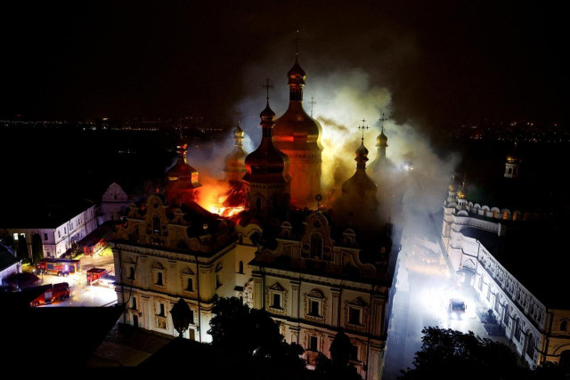 Night view of the Dormition Cathedral at Kyiv Pechersk Lavra engulfed in flames after a Russian attack. Thick smoke rises above the church's golden domes as firefighters work below.