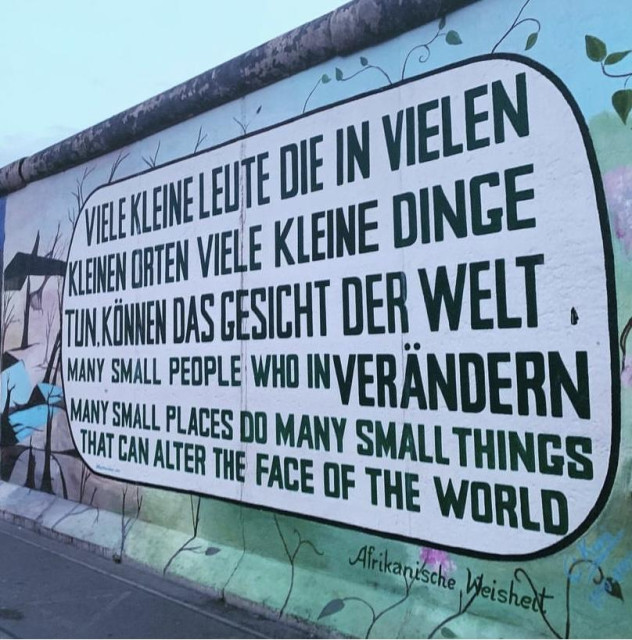 A mural on the Berlin Wall featuring a quote in German and English, emphasizing the impact of small people and places on the world. The text is bold and prominently displayed, with a backdrop of a painted wall.
