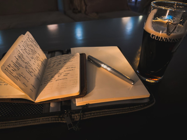 An open notebook filled with handwritten notes rests on a table beside a closed journal and a silver pen. A nearly full pint of dark stout stands to the right, catching the warm ambient light. The scene is dimly lit with soft reflections on the tabletop, creating a calm and contemplative atmosphere suggestive of writing, reflection, or planning in a quiet pub or café during the evening.