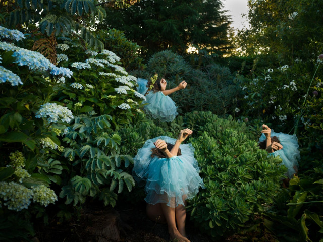 a photograph by Tamara Dean of three individuals sitting in a dense brush, wearing blue tutus around their bodies, only their arms sticking out