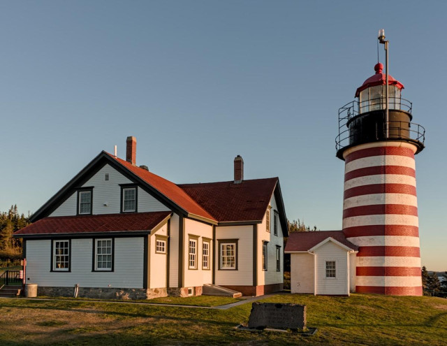 A color landscape photo of a lighthouse and the lighthouse keeper's house. The house is two stories and is painted white with a red roof. There are two sections in the shape of a truncated "L". Each section has a chimney. The light is about twice as tall as the house and stands to its right. It is painted in red and white horizontal stripes. Early morning sunlight shines on the right side of the house and lighthouse tower. A lawn is in the foreground and the sky is clear. 