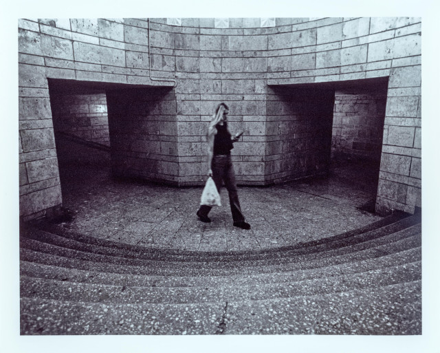 Black-and-white photograph of a woman walking through a curved, tile-clad underground passage, checking her phone while carrying a plastic bag. Two dark, empty doorways flank her on either side, set into a rounded stone wall with a visible pipe running along the top. In the foreground, a set of curved steps descends toward the paved landing where she walks. Shot with a wide-angle lens, giving the architecture a slightly bowed, enveloping perspective. Print shows fine grain and a slight motion blur on the figure.