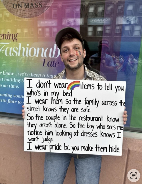 A picture of a man smiling, with a rainbow painted on his cheek and a poster in his hand stating: "I don't wear rainbow items to tell yopu who is in my bed. I wear them so the family across the street feels safe. So the couple in the restaurant know they aren't alone. So the boy who sees me notice him looking at dresses kows I won't judge. I wear pride bc you make them hide."