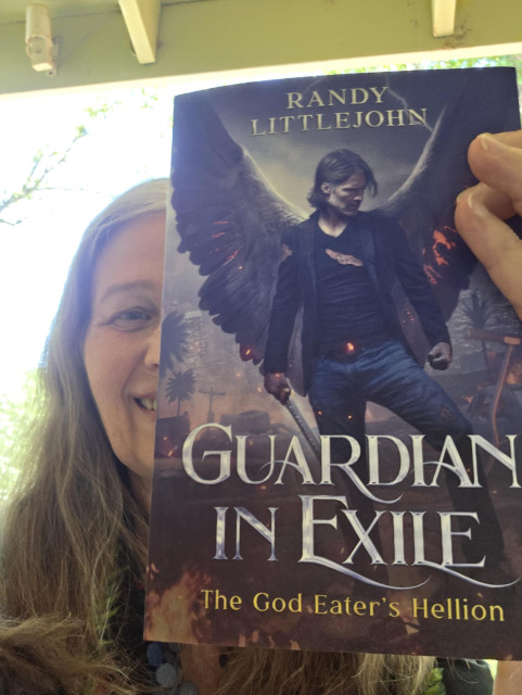 Christy Marx holds up a copy of the Guardian in Exile paperback featuring a man with dark angel wings and a sword, and a stormy Los Angeles in the background. Only the right side of Marx’s smiling face shows from behind the book which is AN AWESOME BOOK. Buy it! Read it! Give it 5-star reviews!