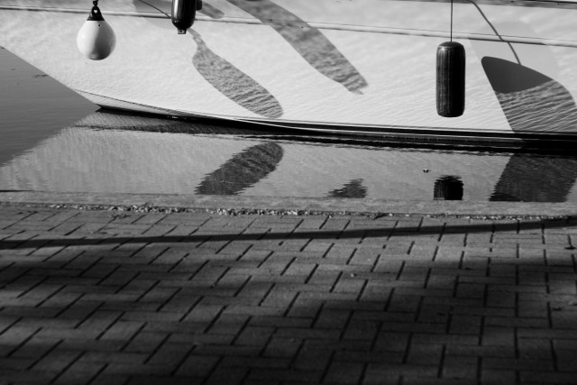 The lower half of a boat with bumpers hanging off of it is seen in this black and white image. Shadows from the bumpers can be seen at different angles as well as the reflection of the boat on the calm water. A cobbled walkway is seen in the foreground also crisscrossed by shadows.