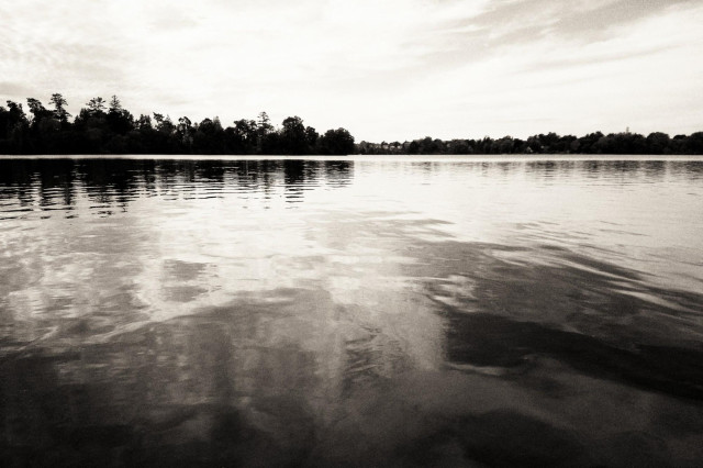 Looking across the calm water of a lake the silhouette of trees along the distant shoreline can be seen. Shapes and patterns can be seen in the calm water from the clouds in the sky as well as the distant trees. This is a black and white image.