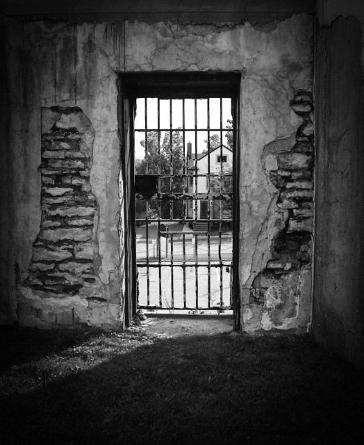 The steel grated door from a old jail cell is seen set in the crumbling wall of what is left of the old jail. Only a few walls of the old building are left set behind a large Provincial courthouse.  This is a black and white image.