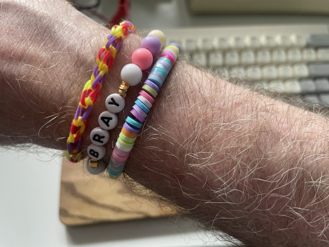 A rather thin, pale, and hairy arm with three hand-made multicolour plastic bracelets.
One has little chains, the other beads and the letters B R A Y and the last litte rings.