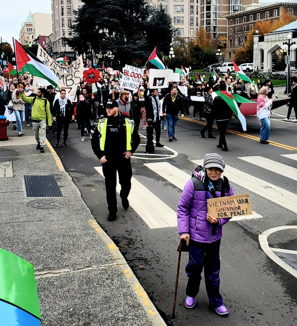 A protest march with people holding Palestinian flags and banners, with me in the foreground holding a cardboard sign that reads "Vietnam War Survivor for Peace!".