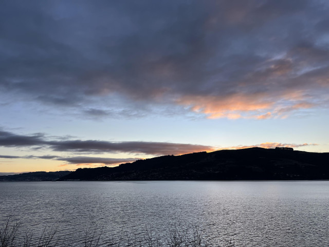 Looking across a rippling Otago Harbour at dusk. There are peachy sunset colours in the partially cloudy sky behind the hill.