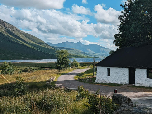 A single track road curves past a whitewashed stone cottage with a dark roof, leading down toward a wide loch flanked by steep green hills. Beyond the loch, mountain ridges recede into hazy blue distance under a sky of stacked clouds. Rough grassland, scrub, and a scatter of trees fill the foreground. 