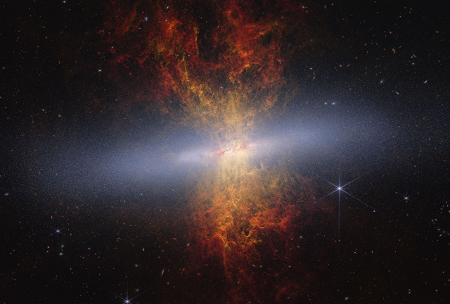 A composite, edge-on view of the starburst galaxy Messier 82 (the Cigar Galaxy), combining data from the James Webb and Hubble space telescopes. The galaxy’s bright, blue-white disk stretches horizontally across the center, glittering with millions of individual stars. Exploding outward from the core—both above and below the disk—is an hourglass-shaped plume of ejected material. Closest to the disk, this plume glows with bright yellow tendrils of ionized hydrogen gas, which gradually give way to expansive, fiery red-orange webs of dust grains tracing the interstellar medium. The vibrant galaxy is set against the deep black background of space, dotted with distant, faint galaxies and a prominent bright star with characteristic eight-pointed diffraction spikes on the right.