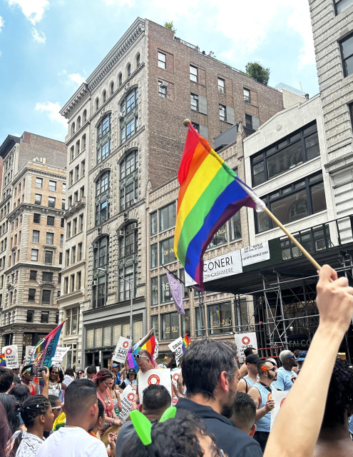 A color photo taken at the NYC pride parade. I couldn't get very high and I'm a little bit in the back. I did crop the photo so that no one was recognizable in the crowd. A girl whose face you cannot see and has black hair wearing green kitty ears, is holding her arm up in the air. She is waving a traditional pride flag that dominates the middle of the photo. The rest of the photo are the heads of people matching in the parade and bits of signs and other flags. Across the street are many old lighter colored buildings. The parade had an amazing energy😊🏳️‍🌈
