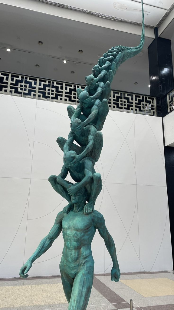 A photograph of a tall, green-patinated bronze sculpture inside a spacious building with white walls. The artwork features a muscular man walking forward at the base. Balanced on his shoulders is another crouched man, who covers the eyes of the man below him. This pattern repeats vertically, with an entire chain of crouched figures stacking on top of one another, each covering the eyes of the person beneath them, curving upward and tapering off into a long, spine-like tail near the ceiling.