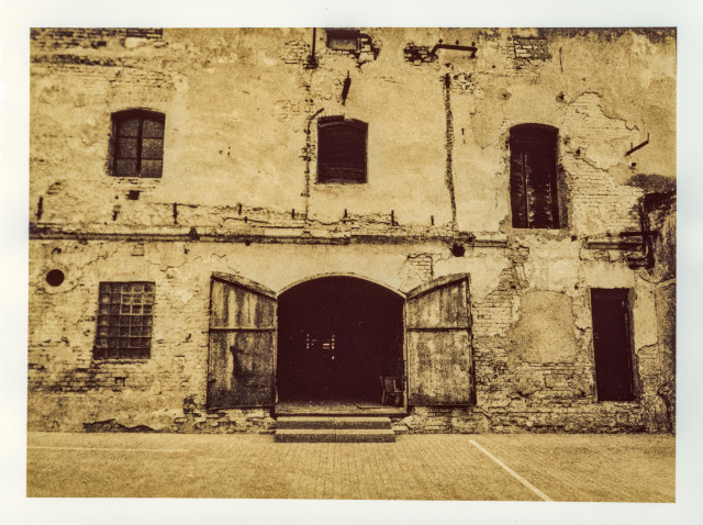 Lith print on Fomatone paper. A crumbling old industrial façade — brick and peeling plaster, arched double doors thrown open onto darkness. A solitary chair sits just inside the threshold. Upper floor punctuated by three shuttered windows. Warm sepia-brown tones with deep, granular shadows characteristic of lith processing.