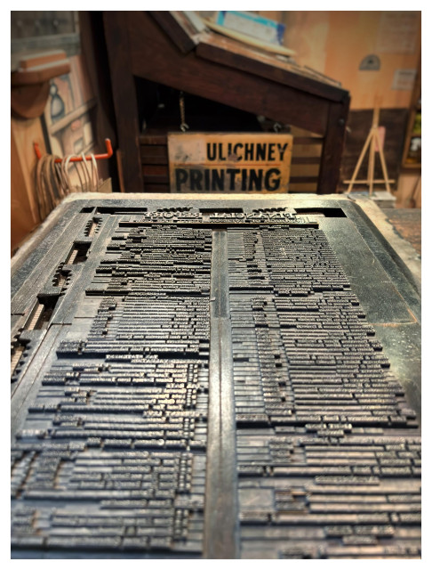 A scene in a vintage printing shop in Pennsylvania. A metal slab holding plates of movable type is viewed from an angle, so that the rows of reversed letters recede from camera like the opening scroll in the Star Wars films. In the background, a sign that reads “ULICHNEY PRINTING” leans against a wooden desk with a slanted surface.