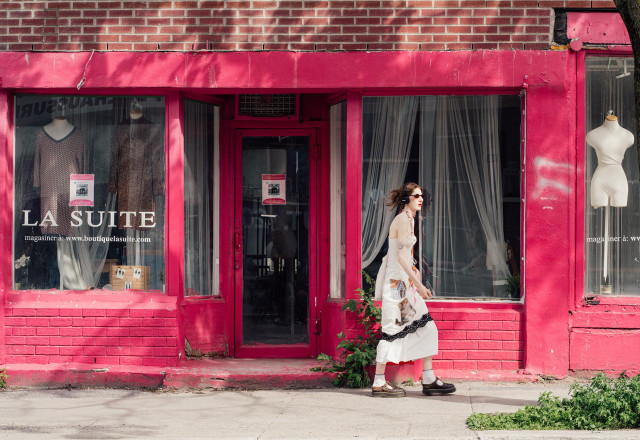 A woman in a white maxi dress passes a brightly pink-painted storefront inhabited only by mannequins in Montreal's Plateau neighbourhood. She's wearing layered jewellery, small black sunglasses, and large earphones; her long dress is adorned with various images: a house, a cat, black lace. 