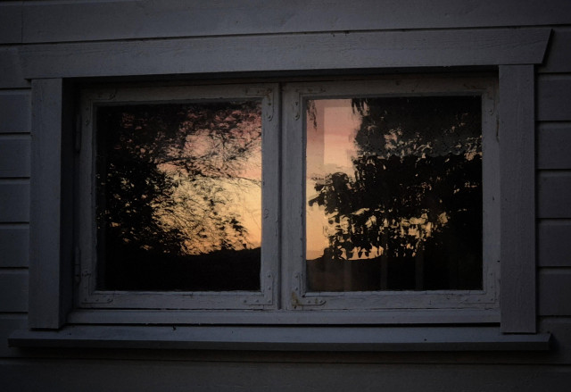 An old, small, white wooden double window, in whose glass a very orange and partly pink sunrise is reflected. The trees appear as black silhouettes against the light. The picture was taken at 3 am