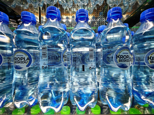 Photo showing rows of plastic  500 ml water bottles with blue caps and labels on a transparent shelf.