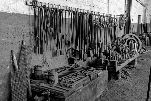 A black and white landscape photo of tools hanging from a rail attached to a wall and lying on a bench. The view is looking at a large concrete brick wall that is painted in a dark color on the bottom and a light color on top. The view is at an angle with the near part of the wall on the left. An iron railing is attached to the wall just above the lower dark section. Hanging from the railing are dozens of various long handled tools used in a forge room (blacksmith). Lying on the bench are 20-25 very large open end wrenches. Most are curved and have been neatly arranged, although, none have been used in many years. The next bench over has a number of other tools, blocks, single jack sludge hammers, and one large iron gear probably three feet in diameter. Another round toothless gear like item is next to the gear and another hangs from the wall. Foot prints are seen on the dirt floor. Everything is very old and dusty.