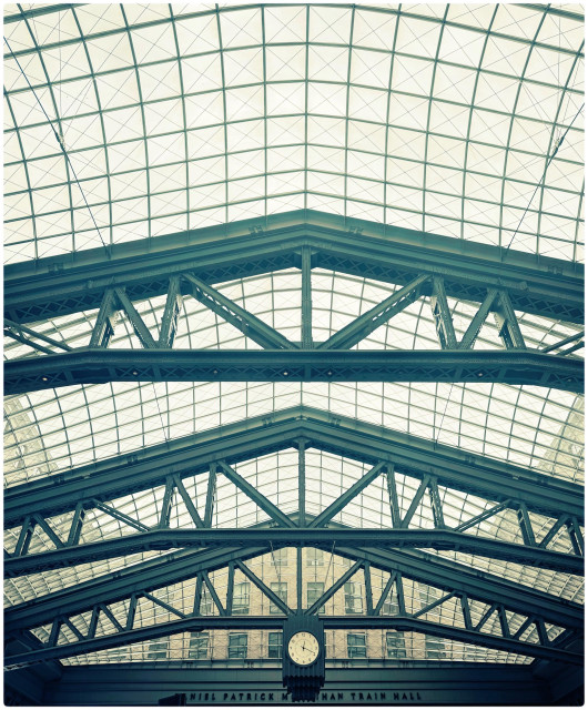 A color photo taken of the window ceiling of Penn Station. The upper 2/3 of the photo is covered in the small square windows of the ceiling. It was a grey day and everything looks very white. The ceiling is being held up in three places by huge steel beams that come to points in the middle and have other steel pieces that make triangles in between another beam going straight across the bottom. The bottom third of the photo shows the third beam through which you can see facing out the window another brick building that looks like it's encased in fog. In the middle of the photo is a round white faced clock surrounded in square black metal on a pole that cannot be seen. Across the bottom is a black metal beam and the words Daniel Patrick Moynahan Train Hall can be seen in tiny letters. 