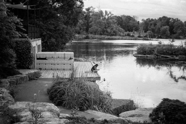 A lone person sits crossed legged at the edge of a dock overlooking a river. Trees can be seen across the water and to the left, large decorative rocks make up part of the path and bank in the foreground. The water is calm and a large stack of floating docks sits behind the person. This is a black and white image.