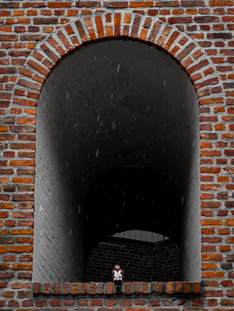A small doll of a man dressed in historical attire sitting on the ledge of a brick window sill, looking out from a dark archway in a brick wall.

The dominant feature is a large, rounded, semi-circular archway built into a textured brick wall. The bricks are predominantly deep red and orange with thick gray mortar lines. 

Faint, vertical streaks of rain are visible falling through the open archway against the dark background.